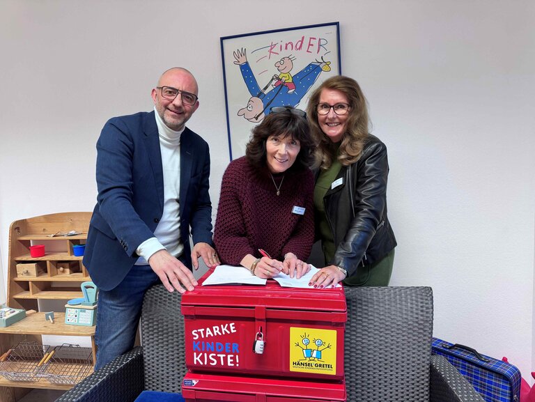 Drei Erwachsene lächeln beim Unterzeichnen eines Dokuments auf einer roten „Starke Kinder Kiste“ der Deutschen Kinderschutzstiftung Hänsel + Gretel, im Hintergrund Kinderposter und Spielzeug.