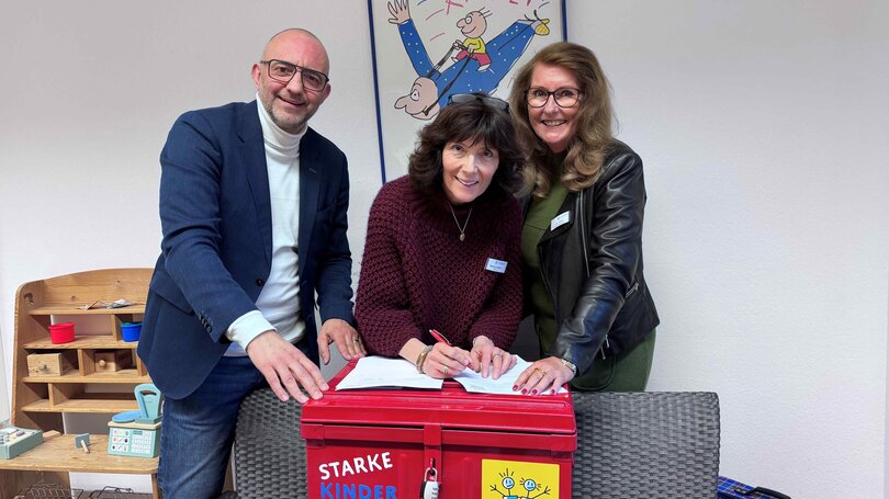 Drei Erwachsene lächeln beim Unterzeichnen eines Dokuments auf einer roten „Starke Kinder Kiste“ der Deutschen Kinderschutzstiftung Hänsel + Gretel, im Hintergrund Kinderposter und Spielzeug.
