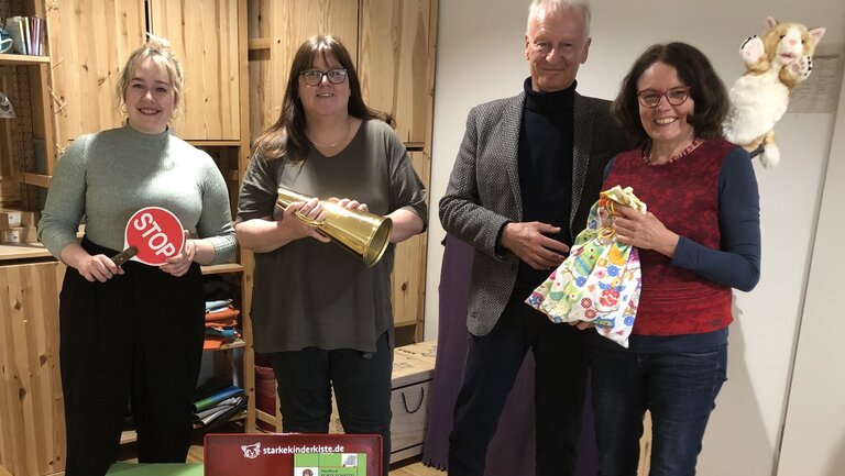Vier Erwachsene stehen vor Holzschränken und halten verschiedene Objekte wie ein STOP‑Schild, eine goldene Tüte und Geschenke; vor ihnen steht die "STARKE KINDER KISTE!".
