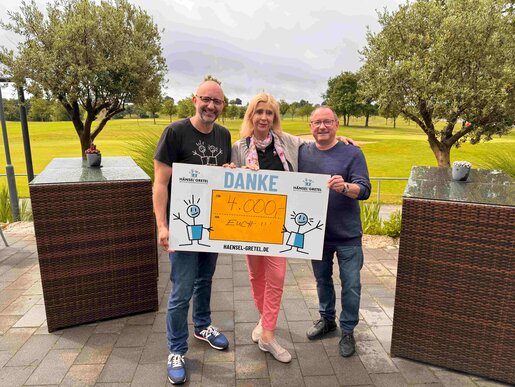 Drei Personen stehen im Freien und halten ein großes Dankeschild mit der Aufschrift „Danke – 4.000 €“ in den Händen; im Hintergrund grüner Golfrasen.