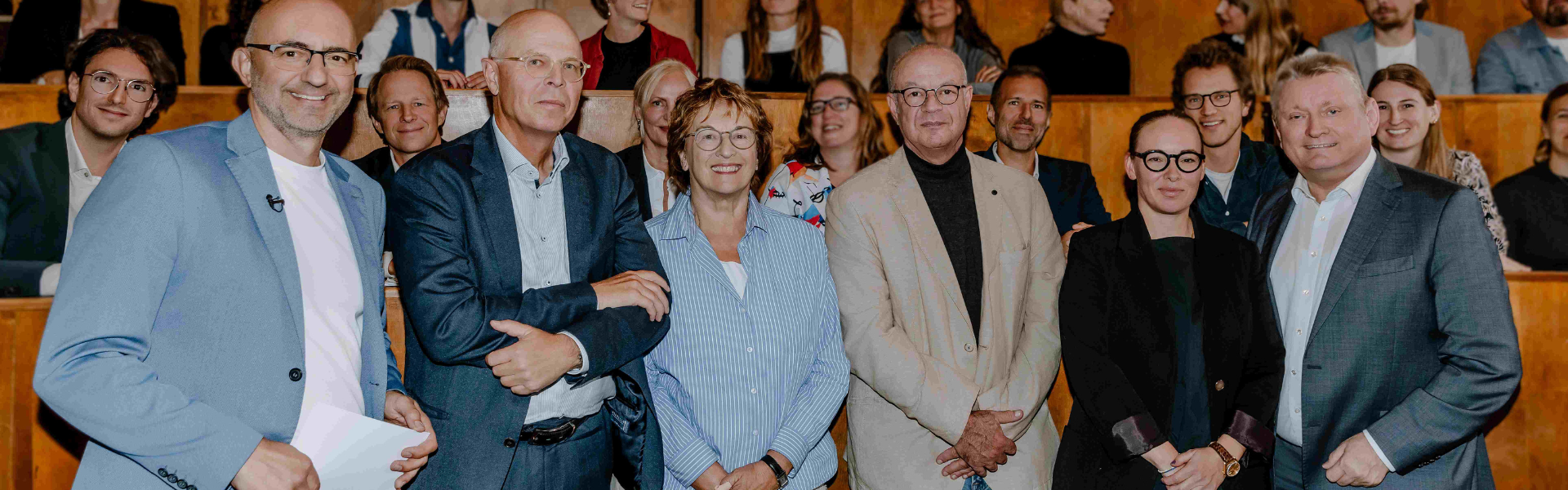 Eine Gruppe von Erwachsenen posiert im Vordergrund eines Hörsaals vor einem Publikum auf ansteigenden Sitzreihen und blickt in die Kamera.