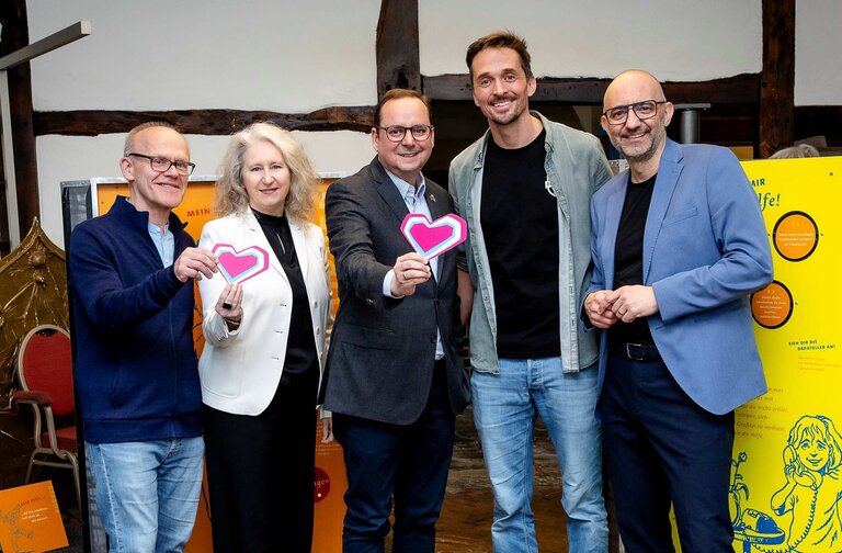 Fünf Erwachsene stehen zur Eröffnung einer Ausstellung zum Thema Kinderschutz zusammen, lächeln und halten herzförmige Karten in die Kamera.