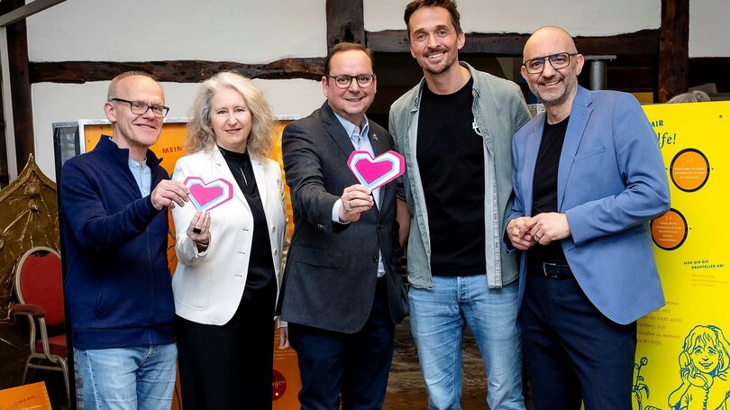 Fünf Erwachsene stehen zur Eröffnung einer Ausstellung zum Thema Kinderschutz zusammen, lächeln und halten herzförmige Karten in die Kamera.