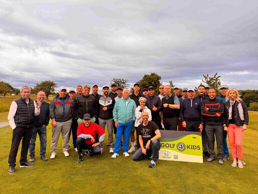 Große Gruppe von Menschen auf einem Golfplatz, die gemeinsam für ein Gruppenfoto posieren; vorne ein Banner mit der Aufschrift „Golf 4 Kids“.