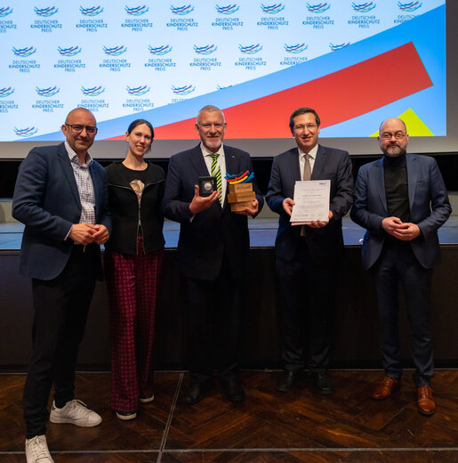 Fünf Personen auf einer Bühne, in der Mitte ein Mann mit Trophäe und Urkunde des Deutschen Kinderschutzpreises.