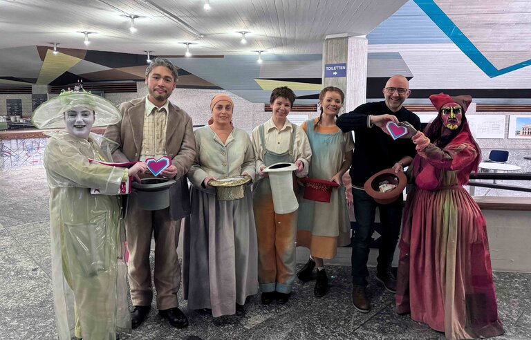 Gruppe von acht Personen in fantasievollen Kostümen aus der Oper „Hänsel und Gretel“ steht nebeneinander in einem Foyer. Sie halten Sammelhüte und Herzsymbole für eine Spendenaktion in den Händen und lächeln in die Kamera.
