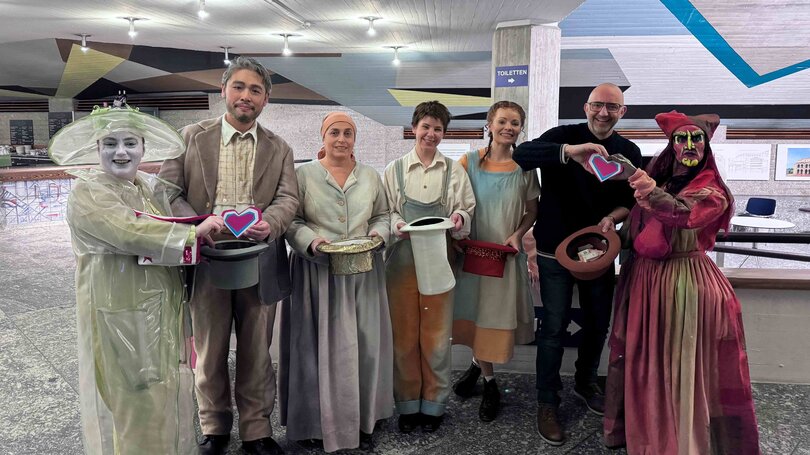 Gruppe von acht Personen in fantasievollen Kostümen aus der Oper „Hänsel und Gretel“ steht nebeneinander in einem Foyer. Sie halten Sammelhüte und Herzsymbole für eine Spendenaktion in den Händen und lächeln in die Kamera.