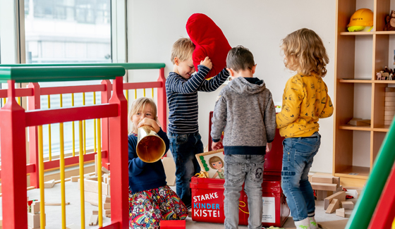 Vier Kinder spielen gemeinsam in einem hellen, freundlichen Raum mit Spielzeug und Holzbausteinen. Eines der Kinder bläst in ein hölzernes Musikinstrument, ein anderes hält ein großes rotes Stoffherz über dem Kopf. Im Hintergrund ist eine rote Kiste mit der Aufschrift „STARKE KINDER KISTE!“ zu sehen. Die Atmosphäre wirkt lebendig und fröhlich.